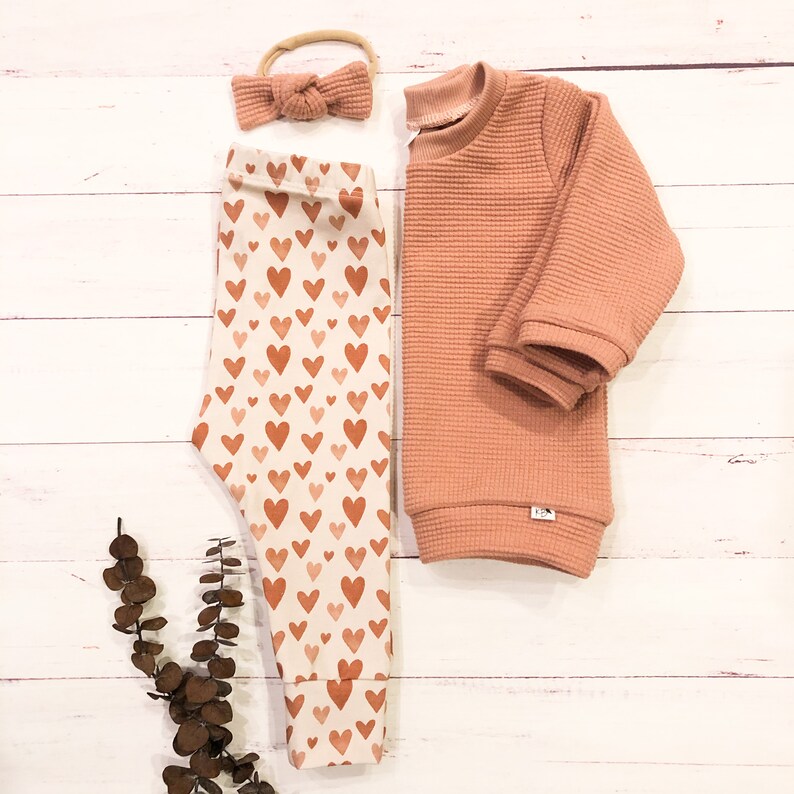 May include: A set of baby clothes featuring a peach-colored waffle knit sweater and white leggings with a brown heart print. A peach-colored headband with a bow is included.