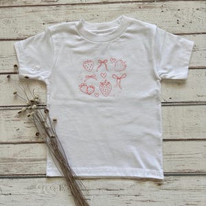 Puede incluir: Camiseta blanca con un diseño de fresas rojas. El diseño incluye fresas, lazos y corazones. La camiseta está hecha de un material suave y tiene mangas cortas y cuello redondo. Adecuada para niños.