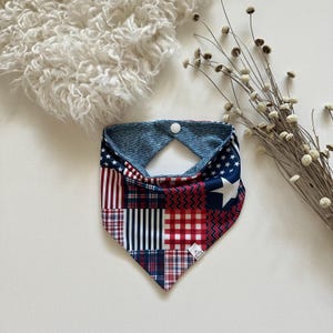 May include: A triangular bandana featuring a patchwork design of red, white, and blue patterns including stars, stripes, plaid, and chevron prints. The accessory has a white snap closure at the top and a blue and white striped fabric on the reverse side.