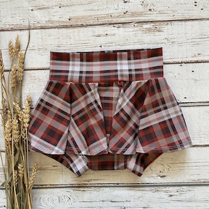 May include: A baby's diaper cover with a red, brown, and white plaid pattern. The diaper cover has a high waist and a ruffled skirt. The fabric is soft and appears to be made of a lightweight material. The background is a light-colored wooden surface.