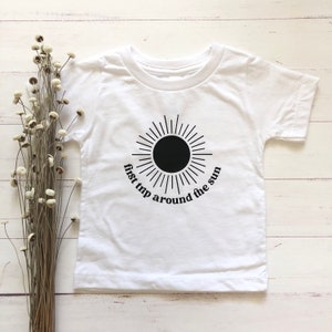 Puede incluir: Camiseta blanca para bebé con un gráfico de sol negro y el texto "first trip around the sun".