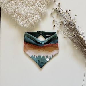 May include: A dog bandana with a forest landscape design. The bandana features a color palette of green, brown, and white, with a button closure at the neck. The design includes a mountain range and trees.