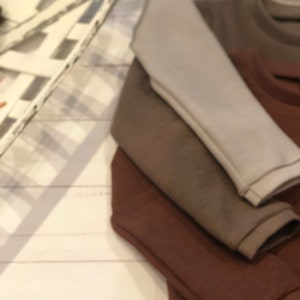 Puede incluir: Una pila de tres camisas de manga larga en tonos grises, marrones y marr&oacute;n oscuro. Las camisas tienen una textura acanalada y est&aacute;n cuidadosamente dobladas, mostrando las mangas. El fondo es una superficie de madera blanca.