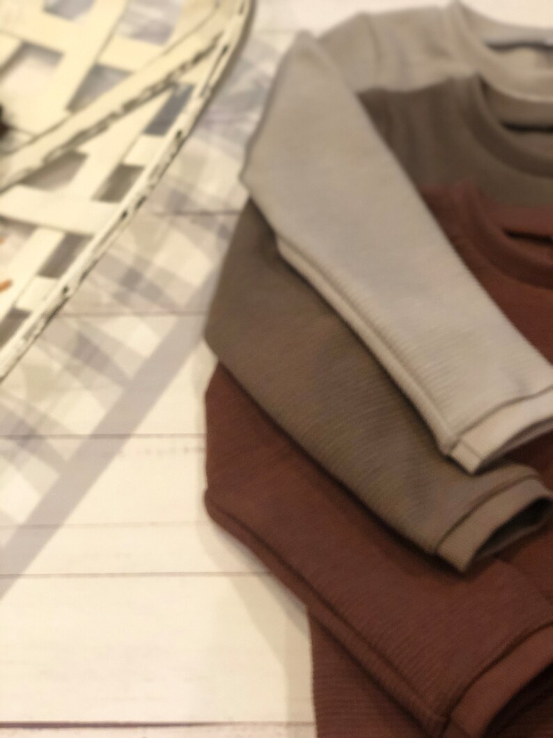 Puede incluir: Una pila de tres camisas de manga larga en tonos grises, marrones y marr&oacute;n oscuro. Las camisas tienen una textura acanalada y est&aacute;n cuidadosamente dobladas, mostrando las mangas. El fondo es una superficie de madera blanca.