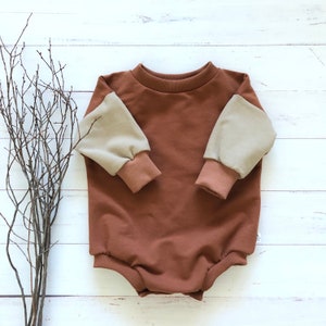 May include: A brown long-sleeved baby onesie with beige sleeves. The onesie has a round neckline and a snap closure at the bottom.