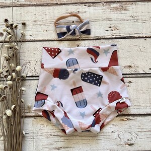 Puede incluir: Un bloomers de bebé con estampado blanco y rojo con un estampado de paletas de hielo rojo, blanco y azul. El bloomers tiene una cintura elástica blanca y una diadema de lazo roja con rayas blancas y azules.