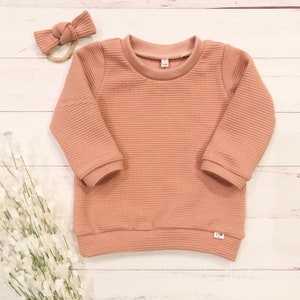 May include: A soft, waffle-knit, long-sleeved sweater in a dusty rose color. The sweater has a crew neckline and a relaxed fit.