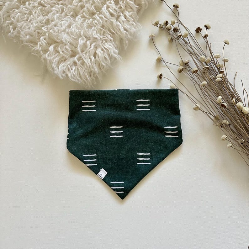 May include: A dark green pet bandana with a white geometric pattern of horizontal lines. The bandana is triangular in shape and has a small white tag on the bottom left corner. The background includes a fluffy white item and dried flowers.