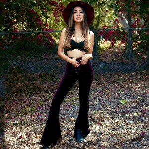 May include: A woman wearing a wide-brimmed burgundy hat, a black lace bralette, and dark purple bell-bottom pants. She is standing in a wooded area with fallen leaves on the ground.