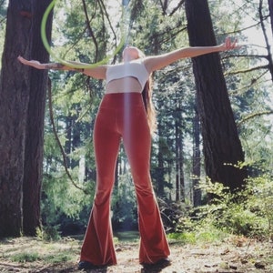 May include: A person wearing a white crop top and red bell bottom pants is holding a green hula hoop above their head in a forest setting.
