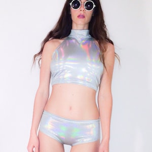 May include: A silver iridescent halter top and matching shorts. The top has a high neckline and the shorts have a low rise. The model is wearing round sunglasses with a white frame.