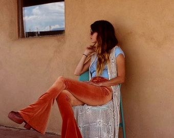 May include: A woman wearing a light blue top with a white crocheted vest and orange bell bottom pants sits on a chair with a white crocheted seat cover. The chair is in front of a tan wall with a window.