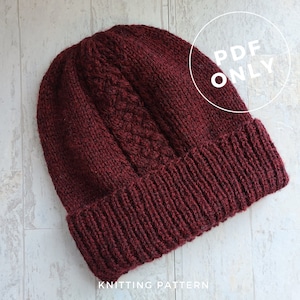 May include: A burgundy knitted hat with a cable knit pattern. The hat has a folded brim and is made with a soft, textured yarn. The image includes the text "PDF ONLY" in a white circle.