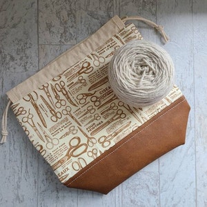 May include: A white fabric drawstring bag with a brown leather bottom. The bag is printed with a pattern of scissors and the text "LAMES RONDES", "CISEAUX", "PLIANTS EN ETUI POUR LA POCHE", "POUR PEINTRES", and "AUX A TOUT FAIRE". A ball of beige yarn is sitting on top of the bag.