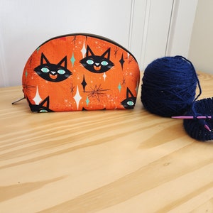 May include: A half-round orange zippered pouch with a black cat pattern and teal stars. A ball of dark blue yarn and a knitting project are on a wooden surface.