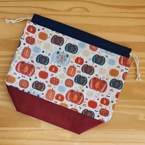 May include: A white drawstring bag with a red and navy blue trim. The bag is decorated with a pattern of colorful pumpkins.