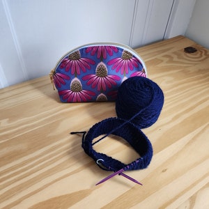May include: A blue and pink floral zippered pouch, a ball of dark blue yarn, and a pair of purple knitting needles on a wooden surface.