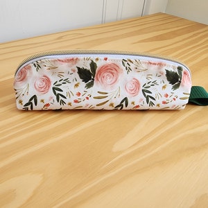May include: A white fabric pencil case with a floral pattern featuring pink roses, green leaves, and small red flowers. The case has a gold zipper and a green fabric strap.