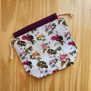 May include: A drawstring bag with a floral rose pattern in shades of pink, burgundy, and yellow on a white background. The bag has a deep purple interior and a cream-colored drawstring. The bag is made of fabric.