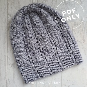 Op de afbeelding: Een grijze gebreide beanie met een geribbelde textuur. De beanie is gemaakt van garen met een subtiel wit vlekje. De afbeelding bevat de tekst "PDF ONLY" in een witte cirkel.