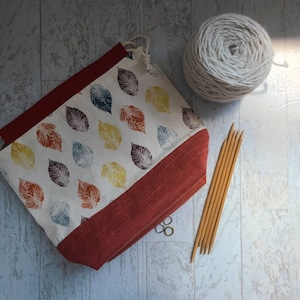 May include: A white drawstring bag with a red border and a leaf pattern. The bag is filled with white yarn and there are wooden knitting needles next to it.