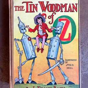 May include: Vintage hardcover book titled "The Tin Woodman of Oz" by L. Frank Baum, illustrated by John R. Neill. The cover features a colorful illustration of the Tin Woodman and a child, with the letter "Z" in a green circle.