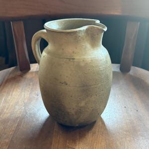 May include: A rustic, beige-colored ceramic pitcher with a rounded body, a curved handle, and a spout. The pitcher has a textured surface and appears to be handmade. It is sitting on a wooden chair.