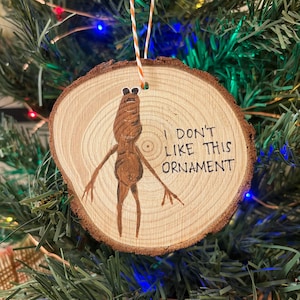 May include: A wooden Christmas tree ornament with a brown painted creature and the text "I DON'T LIKE THIS ORNAMENT". The ornament is round, made from a wood slice, and has a natural wood grain. It hangs from a string.