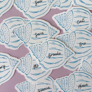 May include: A collection of white paper seashell cutouts featuring blue line art illustrations. Each shell displays a unique name written in black cursive script, including Zoei, Sara, Brooke, Erin, Bianca, and Alexa, arranged in a scattered pattern.