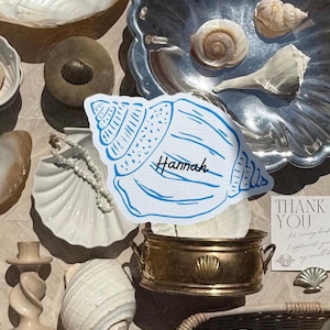 May include: A collection of seashells and decorative items, including a blue and white seashell-shaped place card with the name "Hannah". Other items include a silver tray with seashells, a gold-toned metal container, and a "Thank You" card.