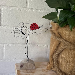 May include: A decorative wire sculpture of a flower and ladybug. The flower is crafted from black wire, with a red ladybug perched on a leaf. The sculpture is mounted on a small wooden base. The ladybug has a red body with black spots.