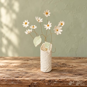 May include: A white cylindrical ceramic vase with a textured floral relief pattern containing a bouquet of handcrafted white daisy flowers with yellow centres, attached to thin gold-toned metal stems with two pale green ceramic leaves.