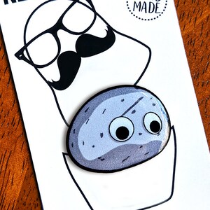 May include: A white card with the text "NEWYORKSHKA" and a cartoon illustration of a face with glasses and a mustache. Below is a blue, oval-shaped pin with googly eyes. The card also has the text "HAND MADE" and a website address.