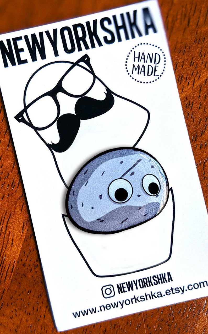 May include: A white card with the text "NEWYORKSHKA" and a cartoon illustration of a face with glasses and a mustache. Below is a blue, oval-shaped pin with googly eyes. The card also has the text "HAND MADE" and a website address.