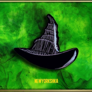 Peut inclure: Épingle noire en forme de chapeau de sorcière avec des détails blancs, sur fond vert et or. L'épingle présente une couronne pointue et un large bord. Le texte "NEWYORKSHKA" est affiché en jaune.