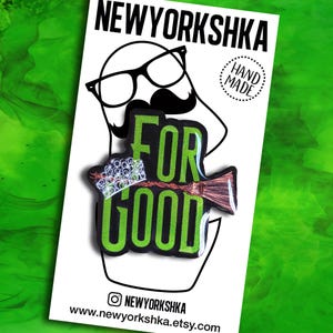 May include: A pin with the words "FOR GOOD" in bright green, with a witch's broom and a crown. The pin is set against a white background with a cartoon face wearing glasses and a mustache. The text "NEWYORKSHKA" is at the top.