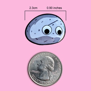 May include: A cartoon rock-shaped sticker with googly eyes, measuring 0.90 inches (2.3 cm) across. The sticker is gray with black accents and a black outline. A U.S. quarter is shown for size comparison. The background is pink.