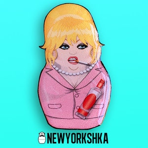 Könnte beinhalten: Handbemalte Matrjoschka-Puppe aus Holz mit blondem Haar, rosa Blazer und einer Flasche Alkohol. Die Puppe hat eine Zigarette im Mund und eine Perlenkette. Die Puppe befindet sich auf einem türkisfarbenen Hintergrund. Der Text "NEWYORKSHKA" steht unten.