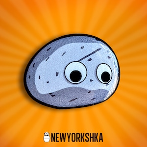 May include: A cartoon rock with googly eyes, set against an orange burst background. The rock is grey with black speckles and a black outline. The logo "NEWYORKSHKA" is at the bottom.