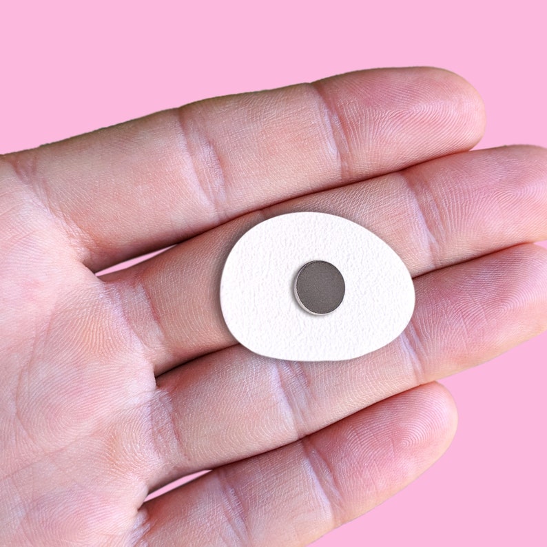 May include: A white, oval-shaped magnet with a textured surface and a small, round, gray magnet in the center. The magnet is held in a person's hand against a pink background.
