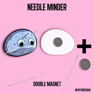 May include: A needle minder set with a cartoon rock design, a white egg-shaped piece, and a double magnet. The text "NEEDLE MINDER" and "DOUBLE MAGNET" are displayed on a pink background.