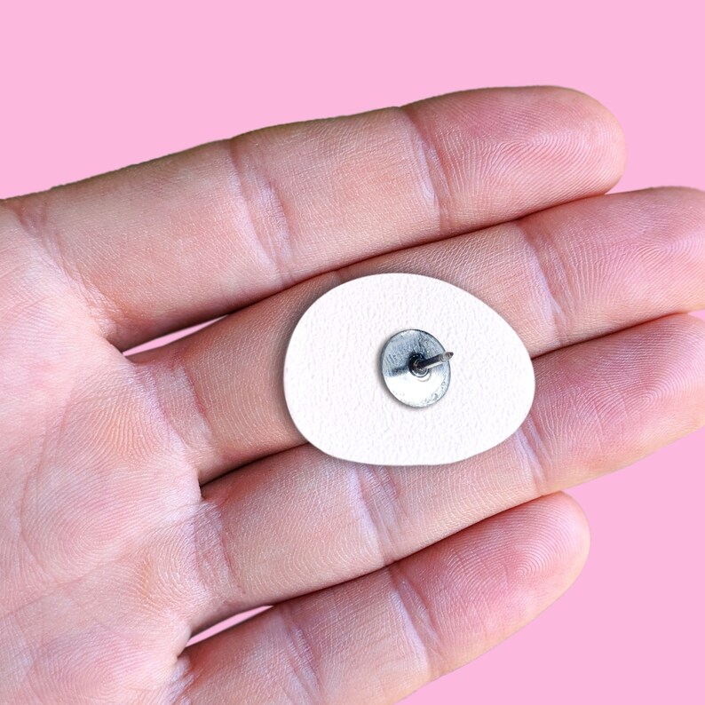 May include: A white, oval-shaped enamel pin with a textured surface. The pin has a silver-colored metal backing with a pin and clasp. The pin is set against a pink background.