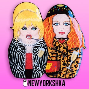 May include: Two wooden matryoshka dolls, one in a zebra-print jacket, the other in a leopard-print coat, each holding a champagne glass. The dolls have painted faces. The text "NEWYORKSHKA" is at the bottom, set against a pink background.