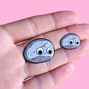 May include: Two cartoon rock-shaped stickers with googly eyes. The rocks are gray with black outlines and white speckles. One is larger than the other. The stickers are held in a hand against a pink background. The stickers are approximately 1.5 inches and 1 inch in length.