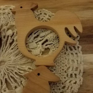 May include: Two wooden rooster-shaped napkin rings. The napkin rings are light brown and have a circular opening in the center. They are placed on a cream-colored, crocheted doily, with a wooden background.