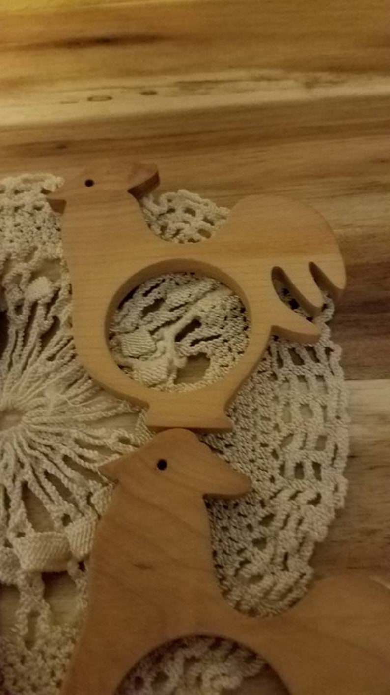 May include: Two wooden rooster-shaped napkin rings. The napkin rings are light brown and have a circular opening in the center. They are placed on a cream-colored, crocheted doily, with a wooden background.