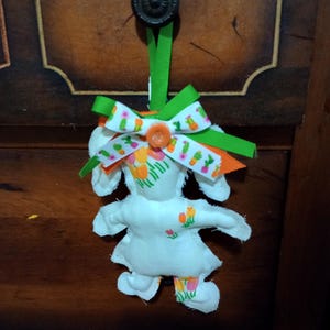 May include: A handmade fabric doll with a white body and floral print dress. The doll has a green ribbon and a decorative bow with cactus and floral patterns. An orange button secures the bow. The doll hangs from a dark brown wooden surface.