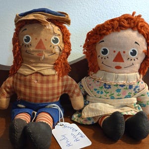 May include: Two vintage rag dolls, one with a plaid shirt and blue pants, the other with a floral dress and white apron. Both dolls have red yarn hair and painted faces with black eyes and a red triangle nose. The doll on the left has a blue and brown hat. A handwritten tag reads "4 inch with pig"