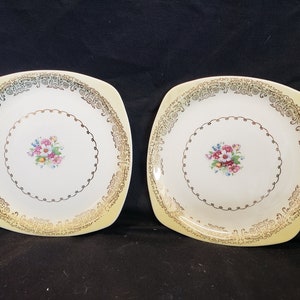 May include: Two square ceramic plates with a gold rim and floral design. The plates are white with a yellow rim and a gold floral design in the center.