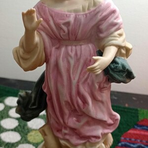May include: A ceramic angel figurine with a pink dress and beige wings. The angel has brown hair, blue eyes, and is holding a dark green object. The figurine is on a white base with a gold trim.